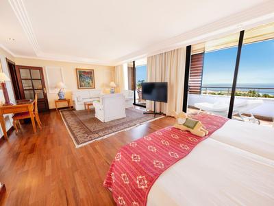 Helles Hotelzimmer mit Doppelbett, Sitzbereich, Schreibtisch und Balkon mit Meerblick.