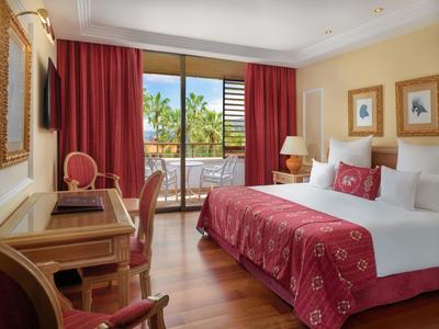 Helles Hotelzimmer mit Doppelbett, rotem Bettüberwurf und Blick auf Terrasse mit Palmen.