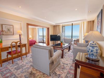 Helles Hotelzimmer mit Sitzbereich, TV und Blick auf das Meer durch große Fenster.