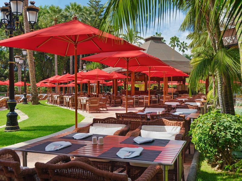 Ristorante all'aperto con ombrelloni rossi e tavoli di legno in un giardino tropicale.