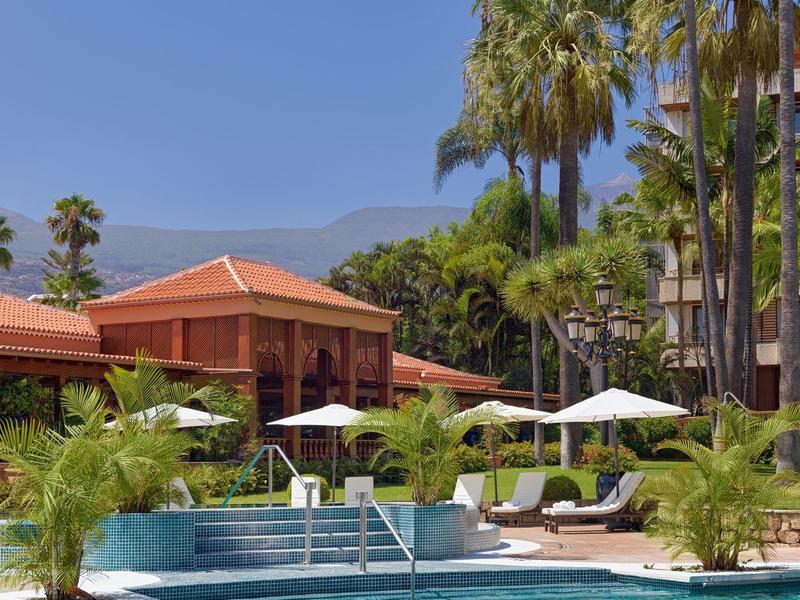 Piscina dall'acqua limpida davanti a palme tropicali e un edificio alberghiero sotto un cielo soleggiato.