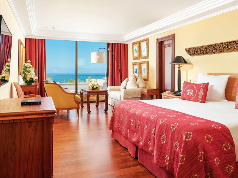 Camera d'hotel luminosa con balcone e vista mare, tende e coperta rosse.