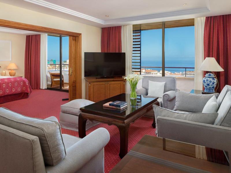 Lounge d'hotel luminosa con area salotto, TV e balcone con vista mare.