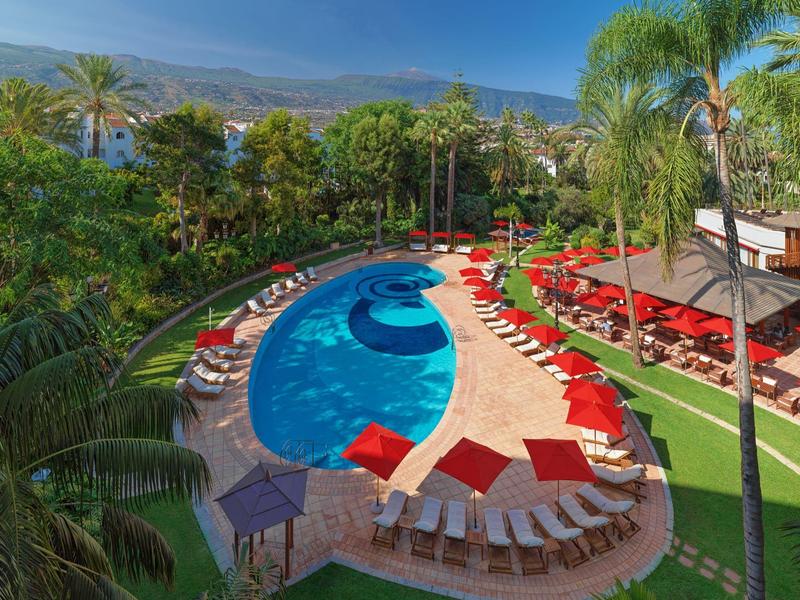 Piscina esterna a forma di fagiolo circondata da lettini rossi e bianchi in un giardino tropicale.