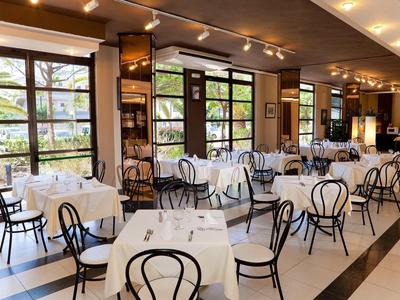 Bright dining room with white tablecloths and black chairs by large windows.