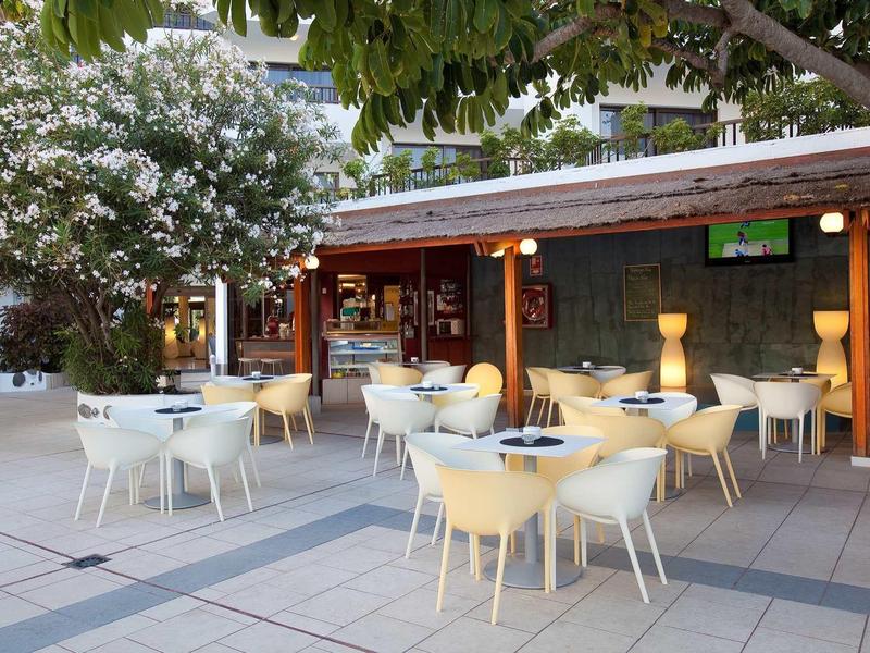Cozy outdoor café area with white tables and chairs under trees and lamps.