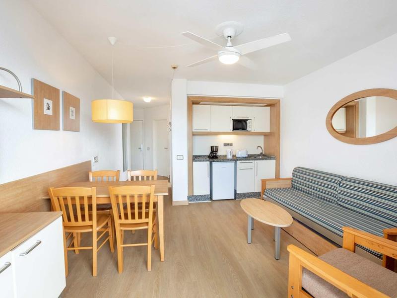 Bright living room with gray sofa, dining table with chairs, and open kitchenette.