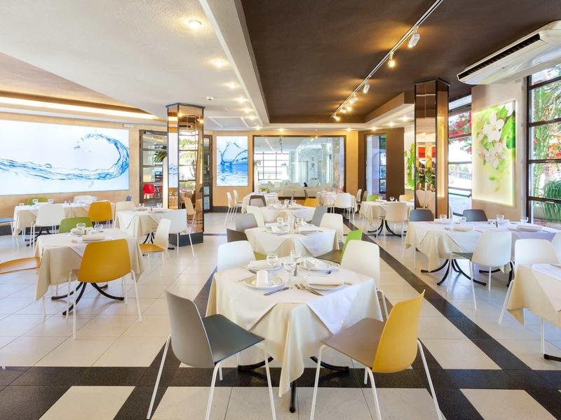 Modern restaurant with white-dressed tables, yellow and gray chairs, and large windows.