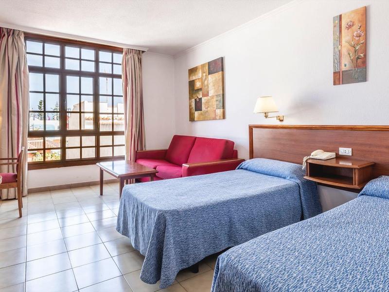 Camera d'hotel luminosa con due letti, TV, scrivania e divano rosso vicino a una grande finestra.