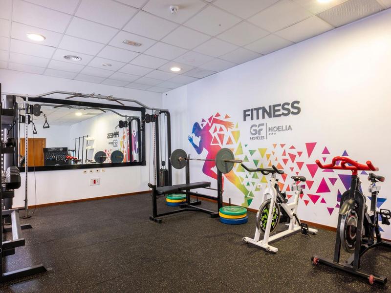 Palestra moderna con pesi, bike da spinning e attrezzature da allenamento davanti a una parete bianca con murales colorato.