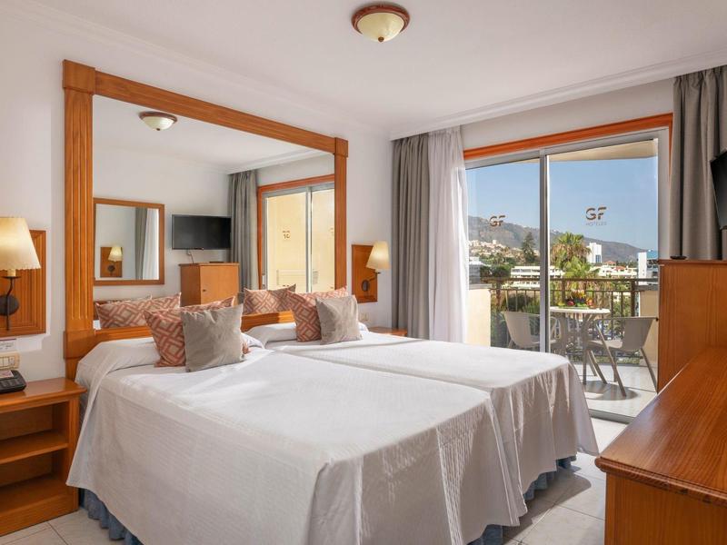 Camera d'hotel luminosa con due letti, mobili in legno e balcone con vista sulle montagne.