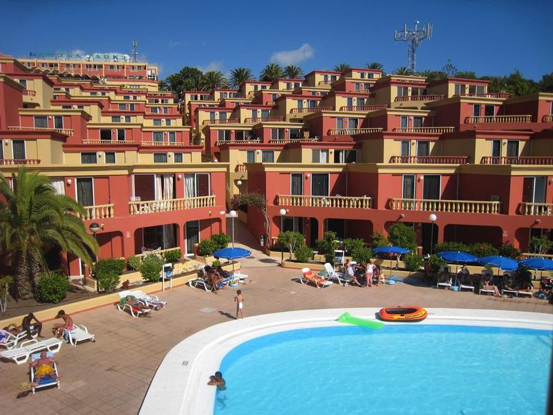 Multi-story hotel complex with outdoor pool, sun loungers, and umbrellas under a blue sky.