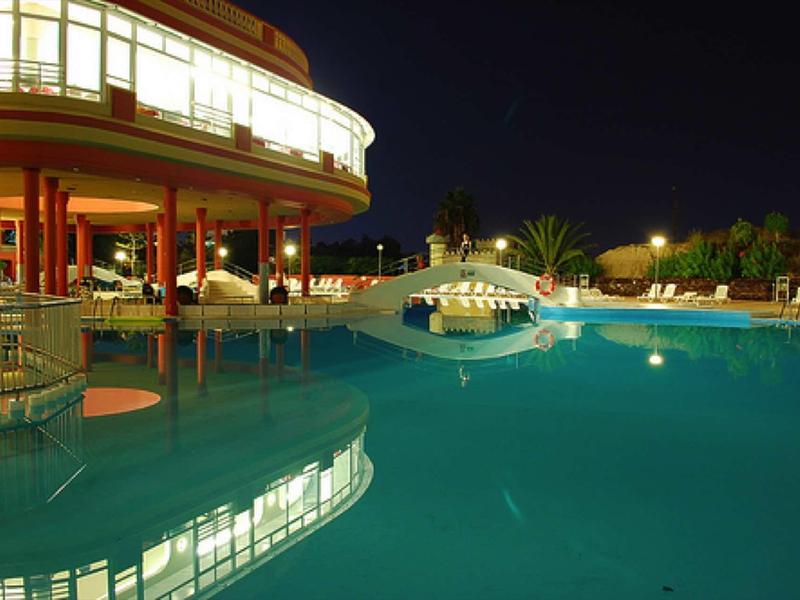 Illuminated swimming pool in front of a hotel building at night with lounge chairs and palm trees.
