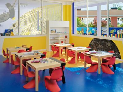 Kindergartenraum mit farbenfrohen Möbeln, kleinen Tischen, roten Stühlen und großer Fensterfront.