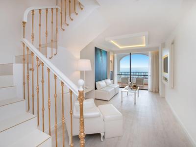 Heller Wohnraum mit weissen Möbeln, Holztreppe, grossem Fenster und Balkon mit Meeresblick.