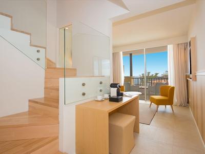 Innenraum mit Holztreppe, kleinem Schreibtisch, gelbem Sessel und Balkon mit Meerblick.