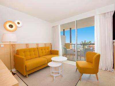 Helles Wohnzimmer mit gelbem Sofa, Sessel, weißen Tischen und Balkon mit Ausblick bei Tageslicht.