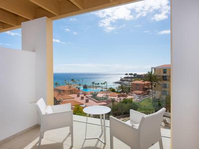 Balkon mit weißen Stühlen und kleinem Tisch, Blick auf Meer, Boote und Häuser unter blauem Himmel.