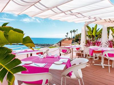Terrassenrestaurant mit rosa Tischen, weißen Stühlen, Palmen und Meerblick unter Sonnensegeln.