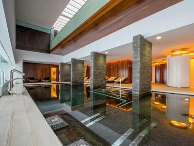 Modernes Hallenbad mit Glasfront, Holzbalken, ruhigem Wasser und gemütlicher Beleuchtung.