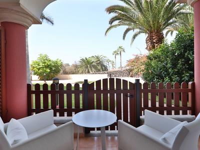 Überdachte Terrasse mit weißen Sesseln, runder Glastisch und Blick auf Garten mit Palmen.