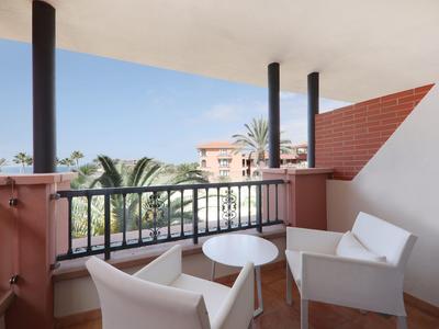 Balkon mit zwei weißen Sesseln, kleinem runden Tisch, schwarzem Geländer und Blick auf Palmen und Gebäude.