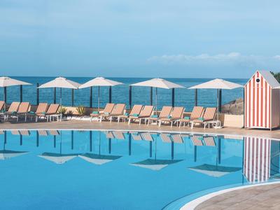 Klare blaue Swimmingpool mit Liegestühlen, weißen Sonnenschirmen und Meer im Hintergrund.