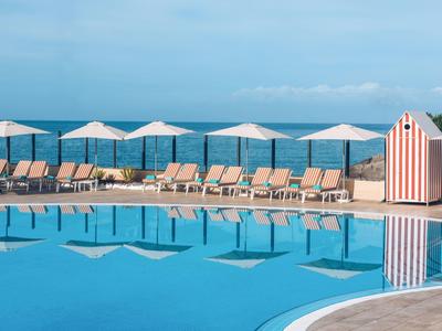 Runder Pool mit Liegestühlen und weißen Sonnenschirmen am Meer, strahlend blauer Himmel.
