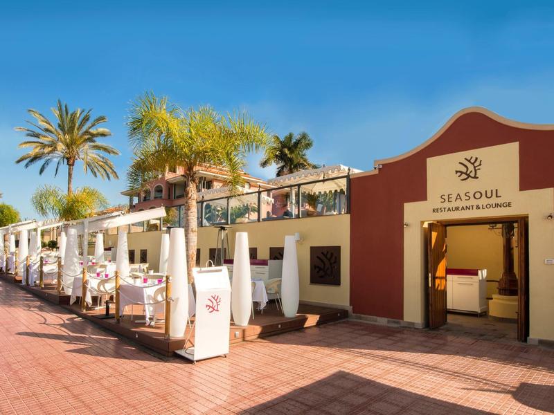Eingang zu einem Restaurant mit Palmen und blauem Himmel, Terrakottaboden und weißer Fassade.