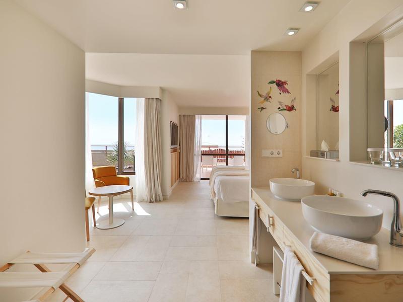 Helles, modernes Hotelzimmer mit Sitzbereich, großem Fenster und offenem Badbereich in warmen Farben.