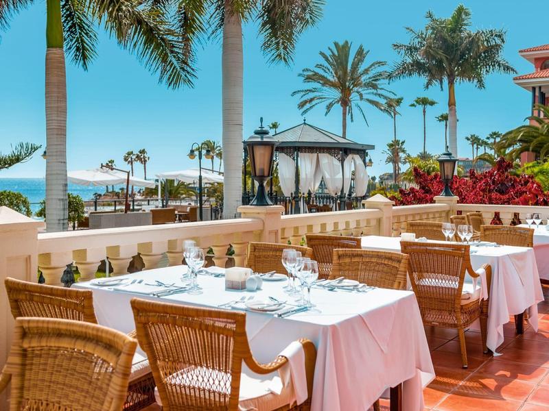 Offenes Restaurant mit gedeckten Tischen, Korbstühlen, Palmen und klarem Himmel im Hintergrund.
