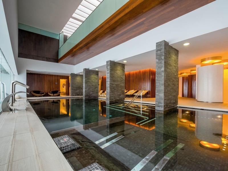 Modernes Hallenbad mit Glasfront, Holzbalken, ruhigem Wasser und gemütlicher Beleuchtung.