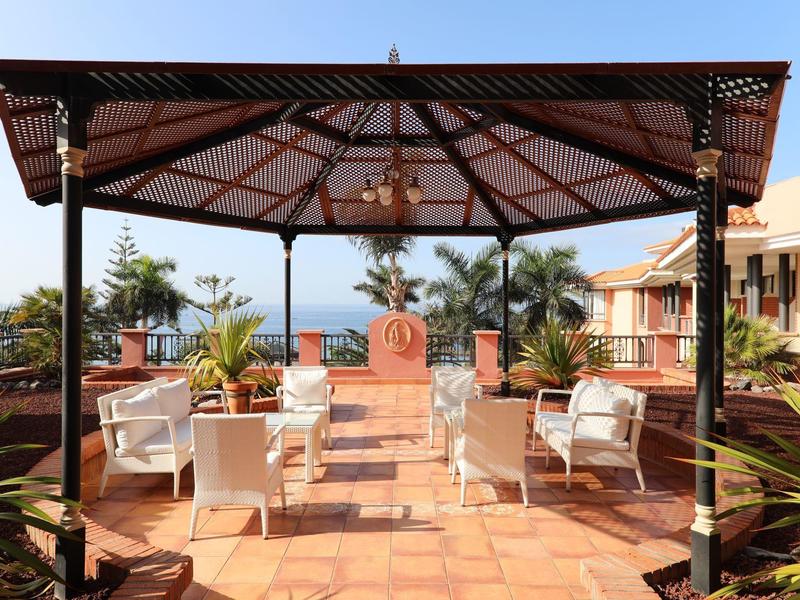 Überdachte Terrasse mit weißen Sitzmöbeln, Pflanzen und Blick auf das Meer bei klarem Himmel.