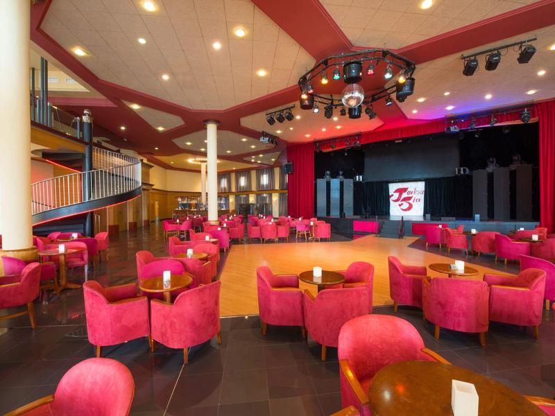 Großer Theatersaal mit rotem Mobiliar, Bühne, Beleuchtung und Holzverkleidung an Säulen.