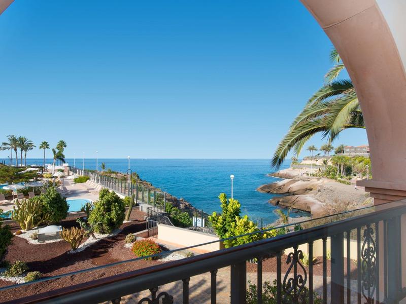 Blick vom Balkon auf blaue See, Palmen, Terrasse mit Pflanzen und klaren Himmel.