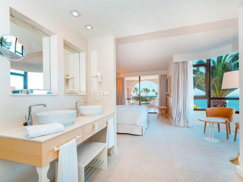 Moderne, helle Hotel-Suite mit Bad, zwei Waschbecken, Schlafbereich und Balkon mit Meerblick.