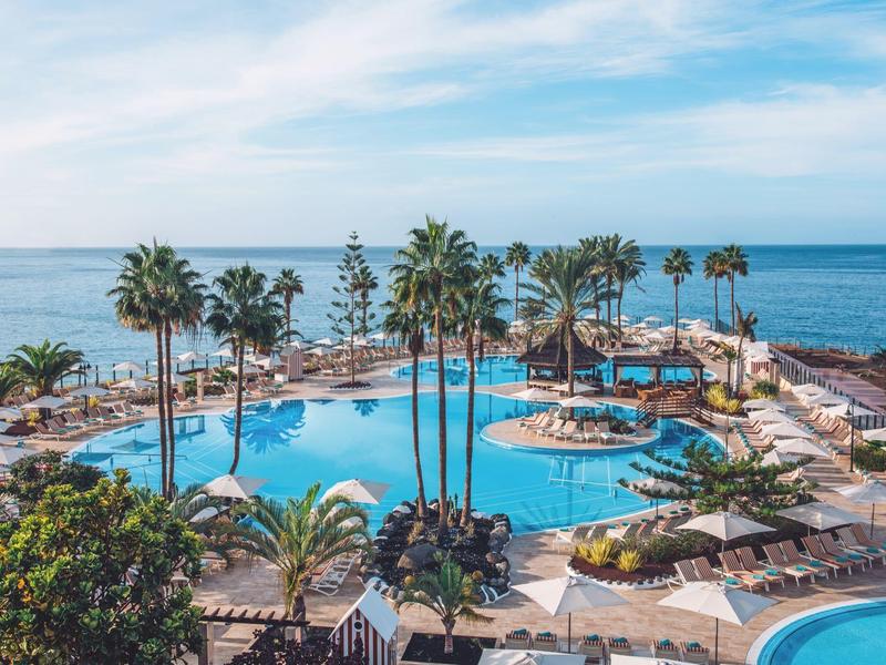 Großer Pool mit Palmen, Sonnenschirmen und Liegestühlen am Meer unter blauem Himmel.