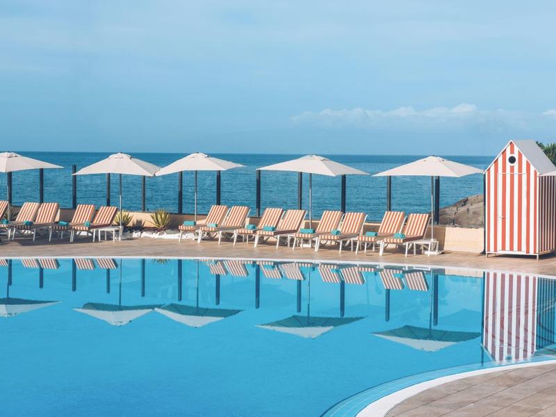 Klare blaue Swimmingpool mit Liegestühlen, weißen Sonnenschirmen und Meer im Hintergrund.