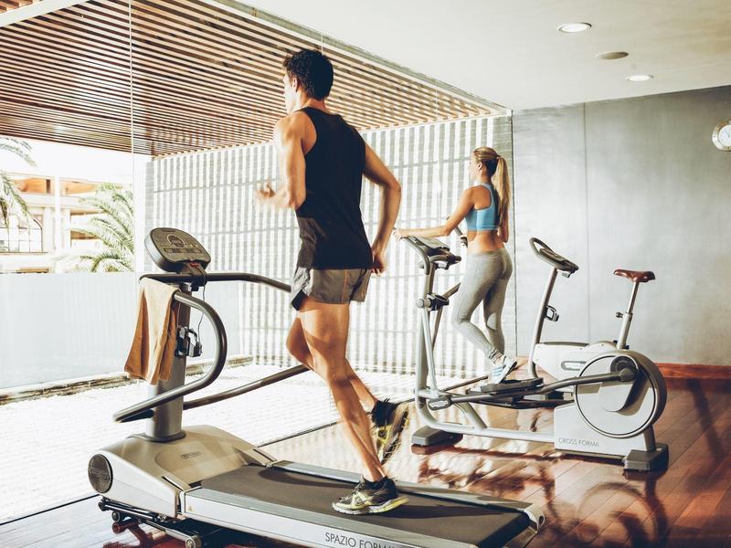 Zwei Personen trainieren in einem hellen Fitnessraum, einer läuft auf dem Laufband, die andere nutzt den Crosstrainer.