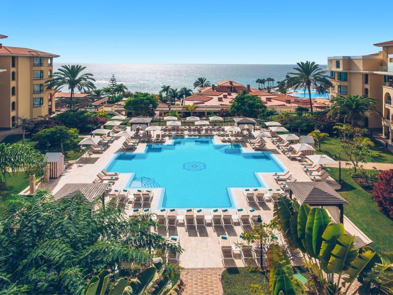 Großer blauer Pool mit Liegestühlen und Palmen zwischen zwei Hotelgebäuden am Meer unter blauem Himmel.
