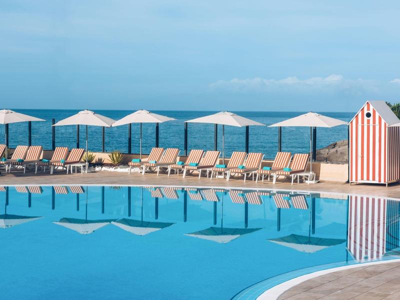 Runder Pool mit Liegestühlen und weißen Sonnenschirmen am Meer, strahlend blauer Himmel.