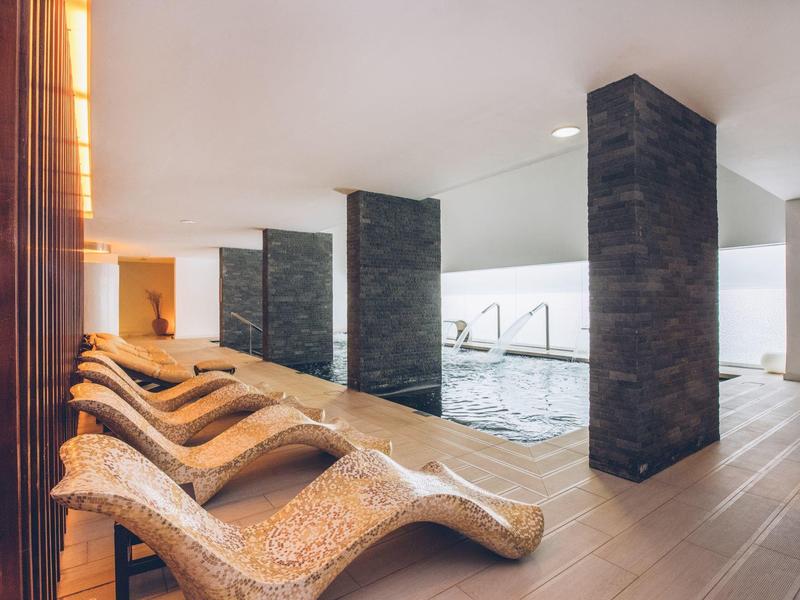 Innenbereich eines Pools mit mehreren Liegen aus Stein und dunklen Säulen zwischen Pool und Sitzbereich.