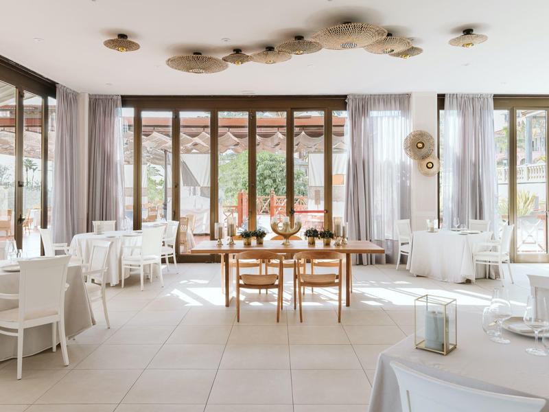 Heller, moderner Speisesaal mit großen Fenstern, weißen Stühlen und Holztisch in der Mitte.