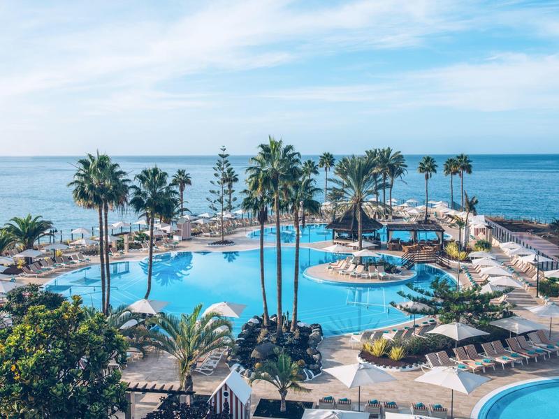 Rundlicher Pool mit Palmen und Liegen, dahinter blauer Ozean unter hellem Himmel.