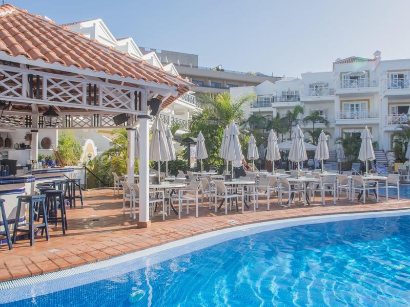 A hotel pool with blue water, lounge chairs, and a covered bar area in sunlight.