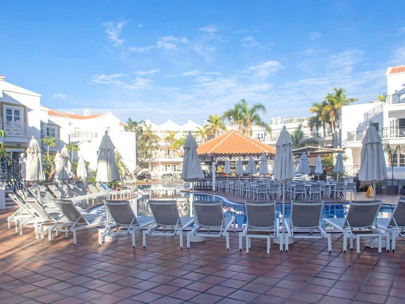 Empty pool area with sun loungers and umbrellas at hotel buildings under a blue sky.