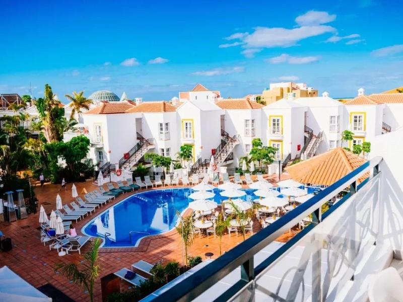 View of a hotel pool surrounded by sun loungers and white buildings under a blue sky.