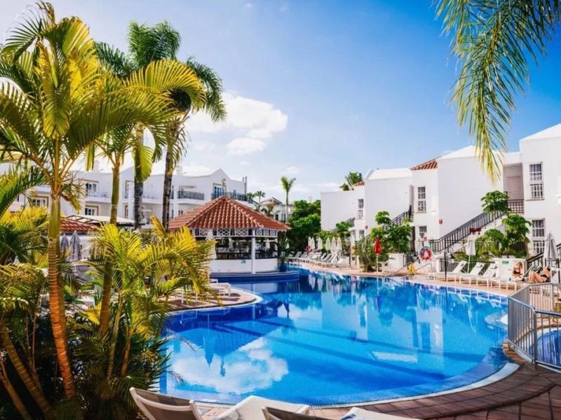 Large pool with palm trees and loungers in front of white hotel buildings on a sunny day.