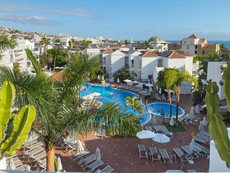 View of a hotel resort with pool, surrounded by palm trees and white buildings under sunny weather.