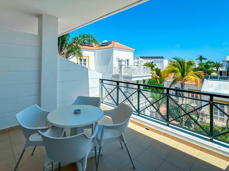 Balcony with table and four chairs, overlooking a palm-lined residential area in sunny weather.